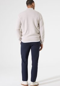 Suéter beige claro con una textura suave, que presenta un cuello. Combinado con pantalones azul marino y zapatillas blancas, visto desde atrás.