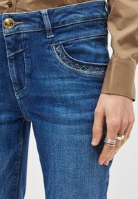 Τζιν παντελόνι σε μεσαίο μπλε χρώμα με κλασική εφαρμογή, που διαθέτει ραμμένες λεπτομέρειες στις τσέπες και διακοσμητικά τρουκς σε χρυσό τόνο κατά μήκος της άκρης της τσέπης.