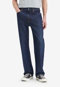 Homme portant un jean droit bleu foncé, une chemise grise claire rentrée et des baskets noires et blanches avec des lacets.