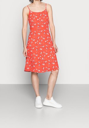 Femme portant une robe orange sans manches avec de petits motifs floraux blancs et des baskets blanches, debout devant un fond uni.