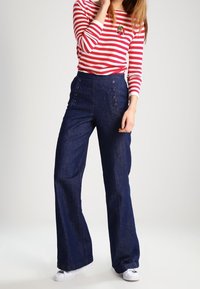 Mørkeblå højtaljede flared jeans med fire dekorative knapper på forsiden, parret med en rød og hvid stribet langærmet skjorte.
