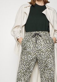 Witte en groene luipaardprint joggingsbroeken met zwarte koord en zakken, gecombineerd met een donkergroen shirt en een lichtbeige buitenlaag.
