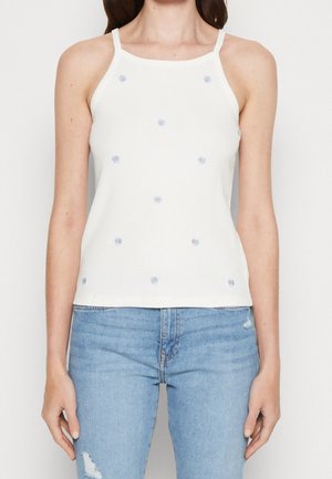 Kobieta w białym prążkowanym topie na ramiączkach z małą niebieską kwiatową haftą i jasnoniebieskich przetartych dżinsach, stojąca na białym tle.