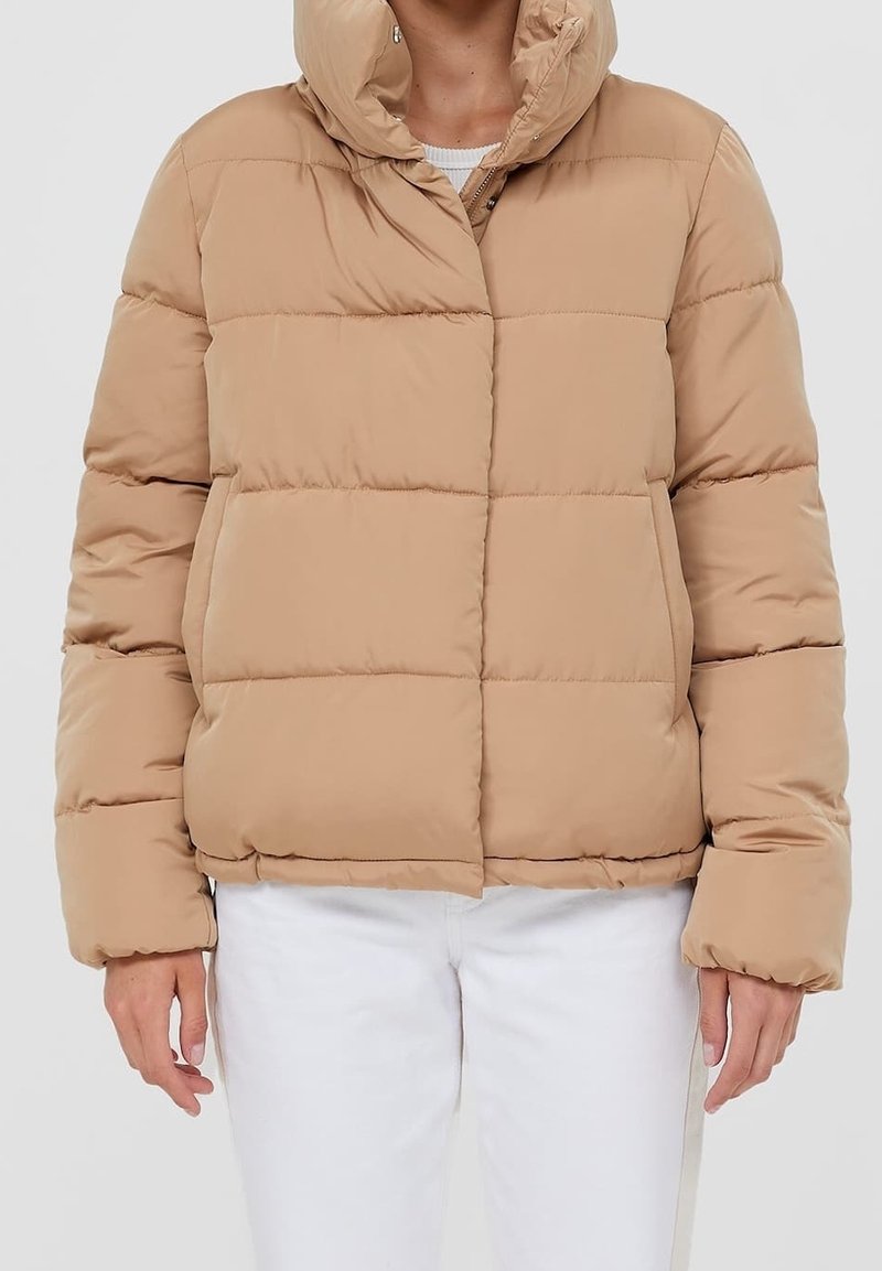 Veste beige matelassée à capuche portée avec un pantalon blanc, vue de face du milieu des cuisses au cou.