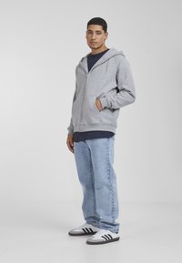 Gray zip-up hoodie with a kangaroo pocket, paired with light blue denim jeans and white sneakers with black stripes and gray accents.