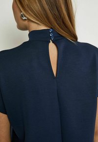 Navy blue top featuring a high collar, short sleeves, and a keyhole back closure with three blue buttons. Soft, textured fabric.
