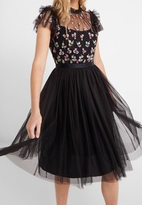 Robe noire avec broderies florales sur un corsage ajusté, manches courtes en tulle transparent à pois et jupe évasée en tulle. Ceinture en satin à la taille.