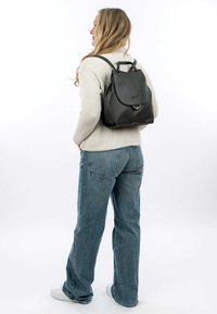 Mochila de cuero negro con una solapa redondeada, un acento de hardware circular, correas resistentes y un acabado texturizado. Llevada en la espalda.