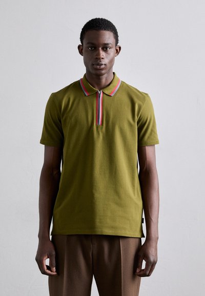Polo shirt vert olive à manches courtes, col zippé, avec des accents rouges et roses. Texture douce, coupe droite, fentes sur les côtés, porté avec un pantalon marron.