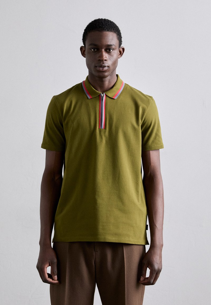 Polo de color verde oliva con mangas cortas, cuello con cremallera y acentos en rojo y rosa. Textura suave, corte recto, con aberturas laterales, combinado con pantalones marrones.