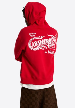 Osoba nosząca jaskrawoczerwoną bluzę z kapturem z białym smokiem i logo „Caballero by Vans” na plecach, w połączeniu z brązowymi, kratkowymi spodniami.