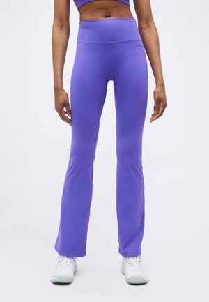 Žena oblečená vo fialových jogových nohaviciach s vysokým pásom a mierne rozšírenými nohavicami, ktoré má obuté s bielymi teniskami, stojaca pred jednoduchým svetlým pozadím.