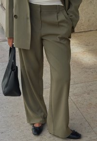 Oseba v olivno zelenih širokih hlačah in usklajenem blazerju, ki drži črn nakupovalni kovček, v črnih baletnih čevljih na marmorni tla.