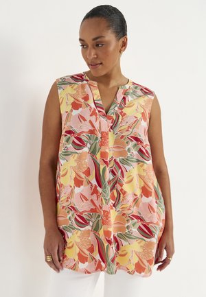 Kobieta ubrana w bezrękawnikową bluzkę w kwiatowy wzór w odcieniach różu, żółci, czerwieni i zieleni, połączoną z białymi spodniami na jednolitym tle.