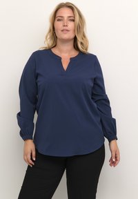 Námořnická blůza s dlouhým rukávem a výstřihem do V, s nařasenými manžetami a volným střihem. Hladká tkanina s jemnou strukturou. Spárována s černými kalhotami.