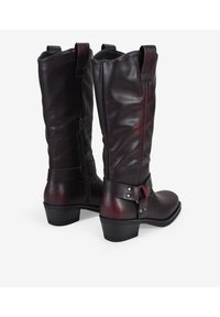 Botas de cuero marrón hasta la rodilla con un acabado texturizado, cierre de cremallera en la parte trasera y correa decorativa con adornos metálicos. Diseño de tacón cuadrado.