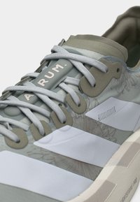 Nahaufnahme eines grauen und weißen Sneakers mit hellgrauen Schnürsenkeln und dem Schriftzug "ARUM." auf der Zunge, versehen mit einem Blumenmuster und einer dicken Sohle.