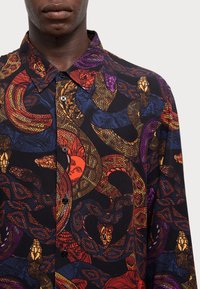 Camicia a maniche corte in tessuto nero con un vivace motivo di serpenti e volti nei toni di rosso, viola e oro. Design con bottoni.