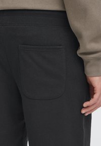 Pantalon de jogging noir en tissu doux, avec une ceinture côtelée et une poche plaquée au dos. La texture semble lisse et confortable.
