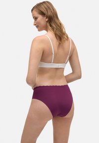 Ropa interior morada de talle alto con una cinturilla festoneada y una textura suave, combinada con un sujetador blanco ajustable, vista desde la parte de atrás.