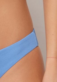 Modré žebrované bikiny, které mají měkkou strukturu a široký pas, částečně viditelné na pozadí v tonálních barvách pleti.