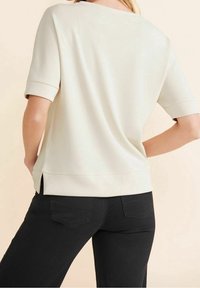 Krémově zbarvený krátký rukáv s kulatým výstřihem, s bočními rozparky a hladkou texturou. Kombinován s černými džínami v pase.