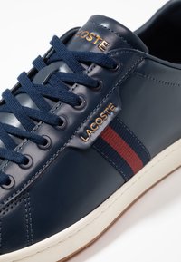 Granatowy skórzany sneaker z fakturowaną cholewką, kontrastującym czerwono-niebieskim paskiem, białą podeszwą oraz złotym logo "Lacoste" po boku.