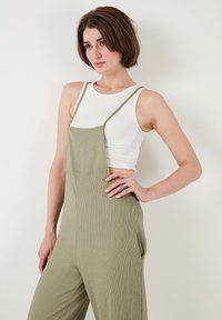 Salopette verde a coste con spalline regolabili e tasche laterali, indossata sopra un top bianco aderente. Tessuto morbido e silhouette rilassata.