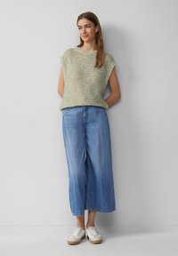 Bezrękawnik w jasnym zielonym i beżowym kolorze, połączony z szerokimi, niebieskimi dżinsami. Model nosi białe sneakersy z brązowymi podeszwami.