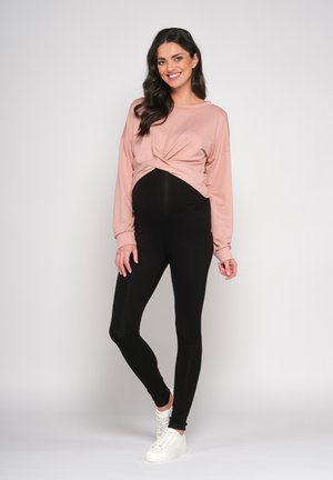 Schwangerschaftsoutfit mit einem hellrosa, langärmeligen Oberteil mit geknoteter Vorderseite und schwarzen Leggings, ergänzt durch weiße Sneakers.