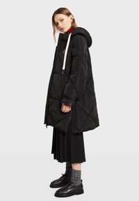 Sort quiltet dynejakke med hætte, med frontlynlås, hvide snore og diagonale syninger. Stylet over et plisseret sort skørt.