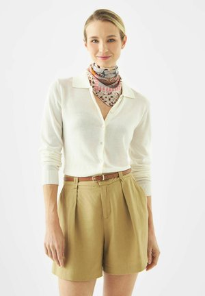 Frau trägt eine weiße, geknöpfte Strickjacke, ein gemustertes Halstuch, beige Shorts mit hoher Taille und braunen Gürtel, steht vor weißem Hintergrund.