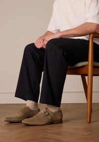 Bruine suède schoenen met een gespdetail, gedragen met licht geprinte sokken, gecombineerd met donkere wijde broekspijpen. Zittend op een houten stoel.