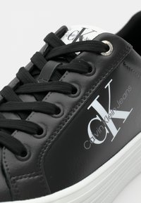 Zwarte synthetische sneaker met veters, voorzien van een wit logo en een getextureerd materiaal. Ronde neus, rubberen zool met witte accentstreep.