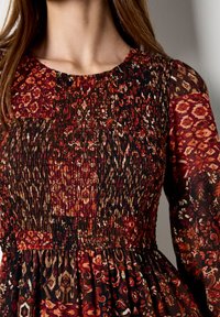 Sukienka w kwiatowy wzór w odcieniach czerwieni, brązu i kremu. Góra jest marszczona, z długimi rękawami i rozkloszowaną spódnicą.