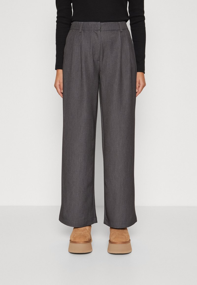 Personne portant un pantalon large gris taille haute, un haut noir à manches longues et des chaussures à plateforme couleur beige, debout devant un fond uni.