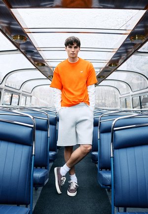 Jonge man in oranjekleurig shirt en grijze korte broek staat met zijn handen in zijn zakken in een lege bus met glazen dak en blauwe stoelen.