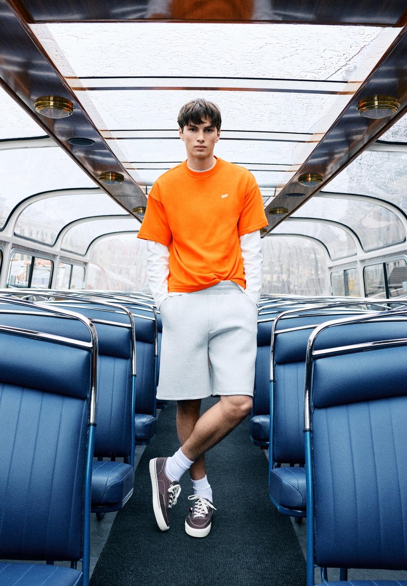 Jeune homme en chemise orange et short gris debout les mains dans les poches à l'intérieur d'un bus vide à toit en verre avec des sièges bleus.