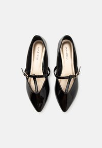 Chaussures en cuir verni noir avec un bout pointu et une fine bride à la cheville, ornées d'une boucle dorée et d'une semelle intérieure nude.