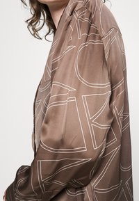 Henkilö, jolla on päällään ruskea satiinipaita, jossa on suuria valkoisia ääriviivoja, paljastaen olkapään ja käsivarren, ja kihara hius näkyy osittain.