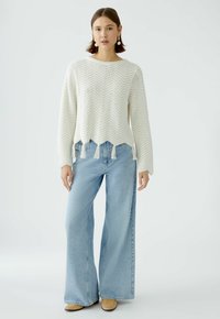 Cremig gestrickter Pullover mit Zickzackmuster, fransigem Saum, kombiniert mit hellblauen, weit geschnittenen Jeans und beige ankle boots. Einfaches, lässiges Design.
