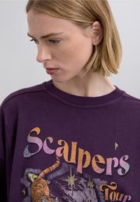 Lilla sweatshirt med tiger-grafik, flerfarvet tekst med teksten "Scalpers Tour", metalliske accenter og en blød tekstur.