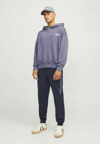 Sudadera con capucha morada y bolsillo frontal, emparejada con pantalones de chándal azul marino que tienen un estampado de logo. El modelo lleva una gorra azul clara y zapatillas.