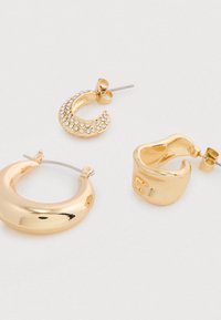 Tres pares de pendientes de aro en tono dorado, uno de ellos con rhinestones, dispuestos sobre un fondo blanco liso.