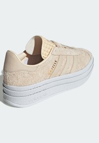 Beige suede sneaker with three textured stripes, rubber white sole, and woven accent on the heel. Features "Gazelle" branding in gold.
