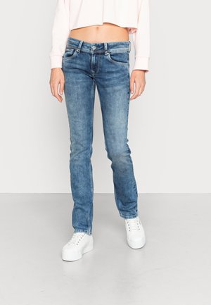 Femme portant un jean bleu slim taille mi-haute, un sweat-shirt court rose clair et des baskets blanches à plateforme, debout sur un sol gris clair uni.
