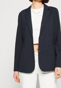 Marineblazer gemaakt van lichtgewicht stof met twee voorkanten zakken, notched revers en twee knopen, gecombineerd met een wit cropped top.