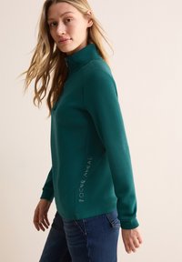 Türkiser Sweatshirt mit hohem Kragen, tailliertem Design und seitlichem Textdetail. Kombiniert mit dunkelblauen Jeans. Glatte Textur, lässiger Stil.
