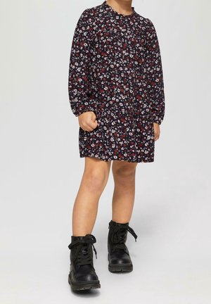 Niña vestida con un vestido floral de mangas largas con flores negras, rojas y blancas, combinado con botas de tobillo negras con cordones, de pie sobre un fondo claro.