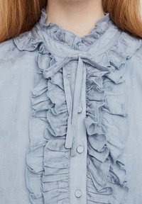Blusa azzurro chiaro con dettagli arricciati, colletto rotondo e una cravatta al collo. Tessuto liscio con una leggera trama e chiusura a bottoni.